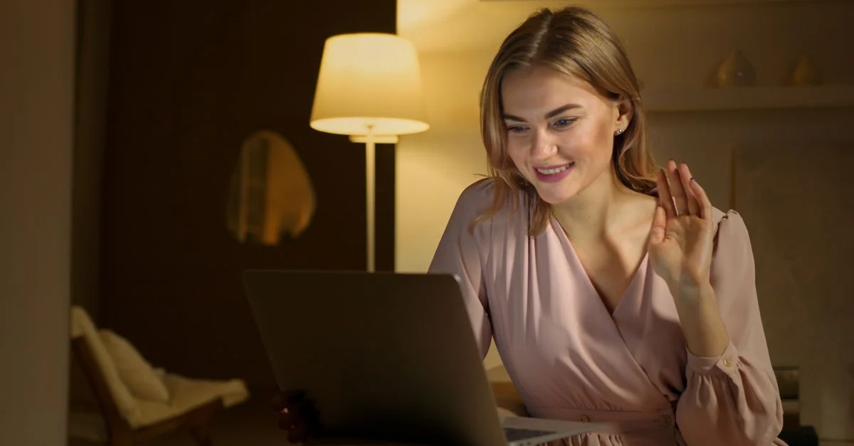 Femme souriante et faisant signe pendant un appel vidéo, utilisant un ordinateur portable dans un cadre intérieur confortable.