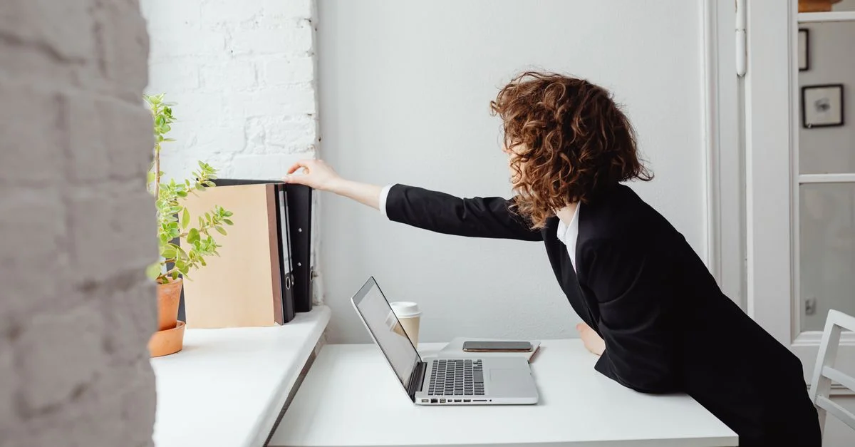 Femme d'affaires atteignant des dossiers sur une étagère tout en travaillant sur un ordinateur portable dans un bureau lumineux.