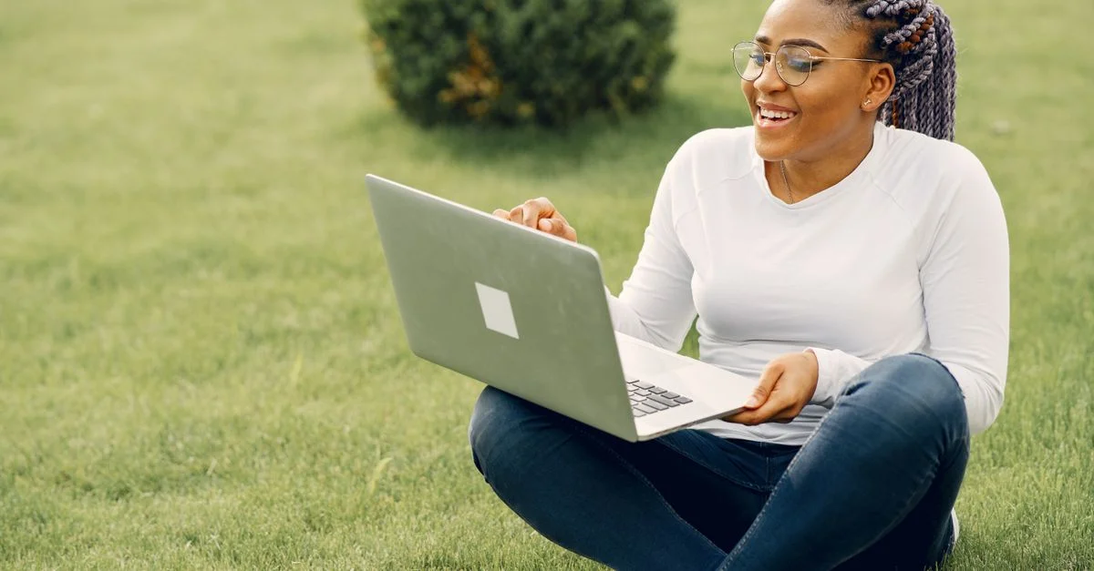 Femme souriante avec des cheveux tressés travaillant sur un ordinateur portable en plein air au soleil.