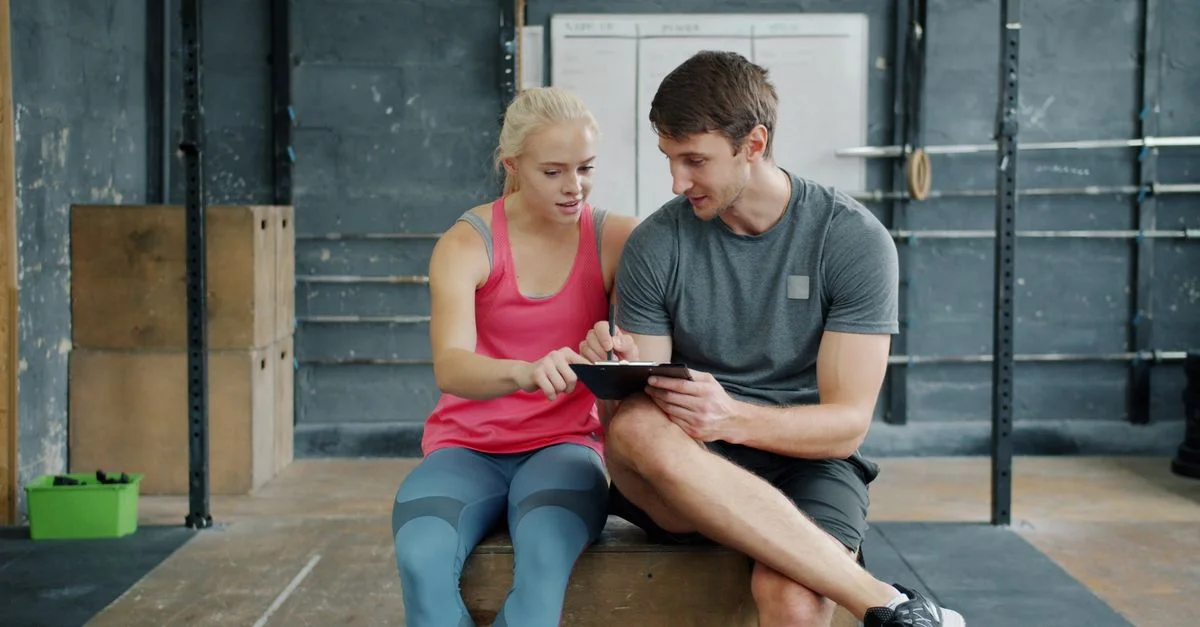 Un homme et une femme en tenue de sport consultent leur programme d'entraînement sur une tablette à l'intérieur d'une salle de sport.
