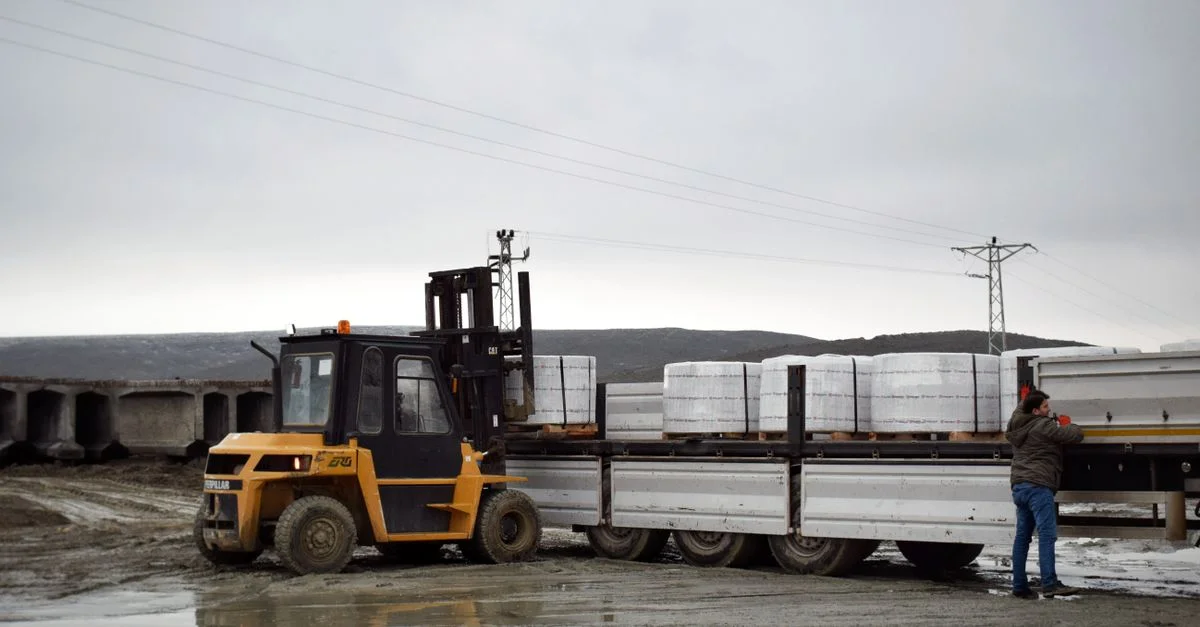 Un chariot élévateur charge des marchandises sur un camion dans une zone logistique boueuse avec un travailleur qui aide.
