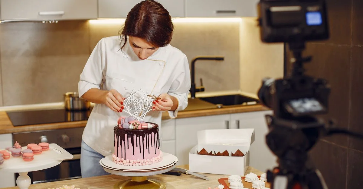 Une femme chef décore un gâteau au chocolat avec un glaçage en coulée dans une cuisine moderne, entourée d'ustensiles de pâtisserie et d'ingrédients.