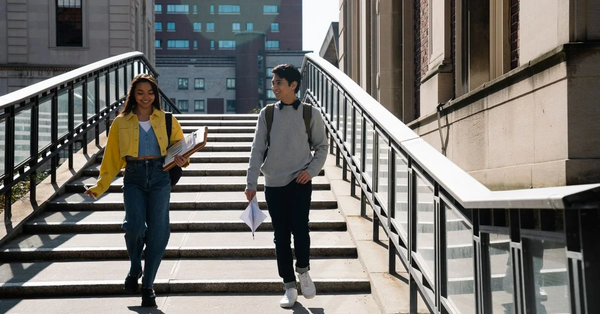 Deux étudiants universitaires descendent un escalier, portant des documents et souriant pendant la journée.