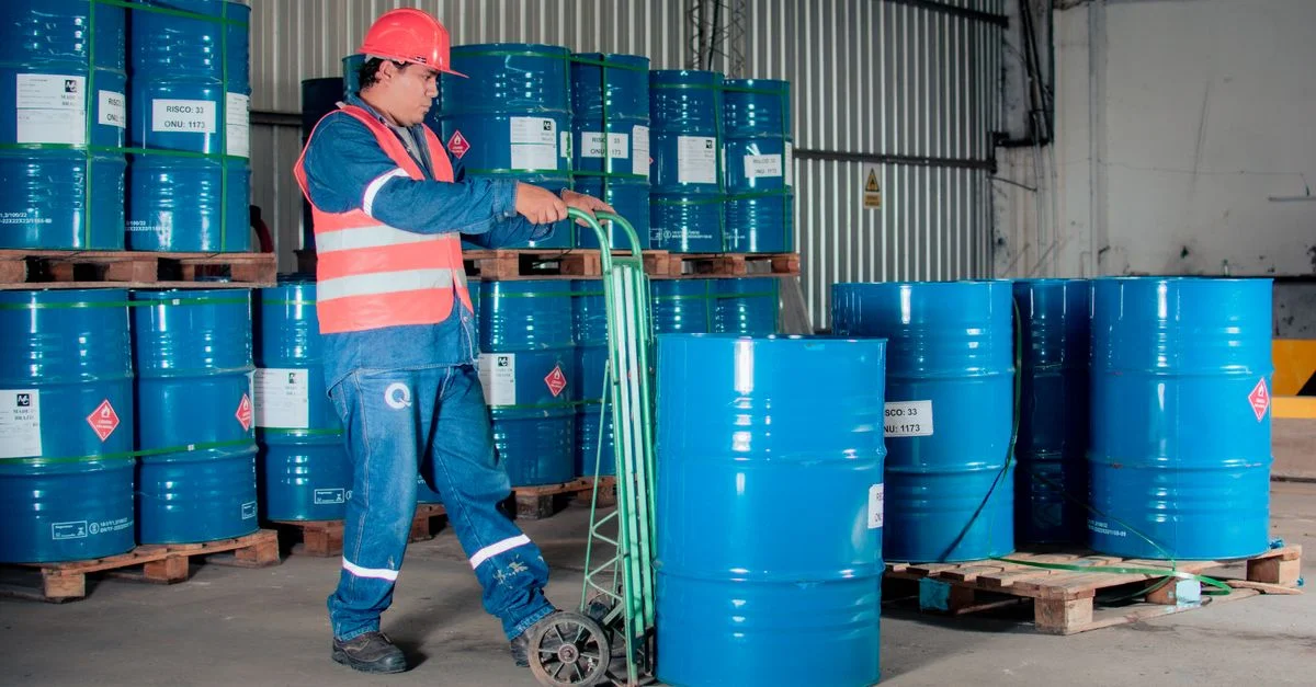 Travailleur industriel avec un diable déplaçant des fûts bleus dans un environnement d'entrepôt.