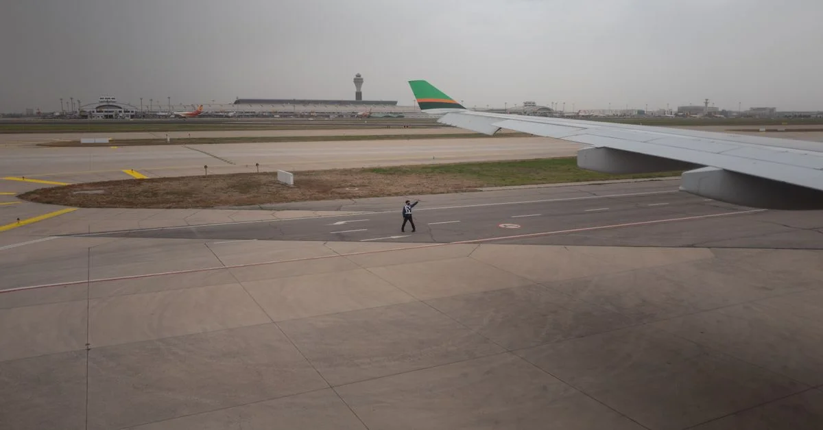 Vue d'une piste d'aéroport avec une aile d'avion, une tour de contrôle et des employés au sol guidant par un jour nuageux.