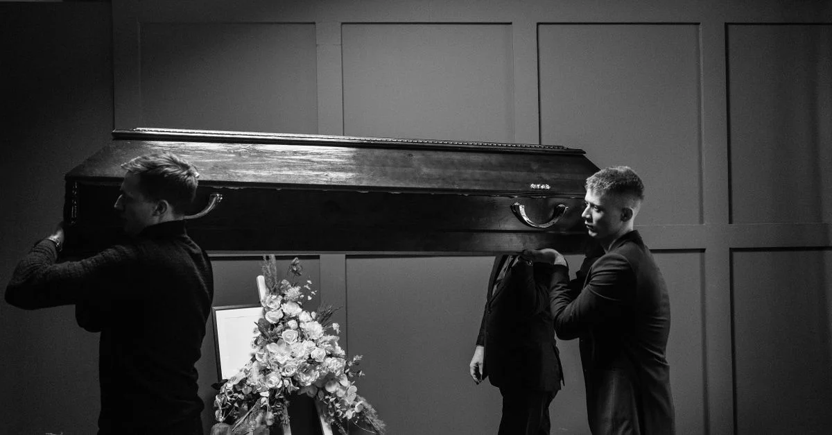 Des hommes portant un cercueil en bois dans un cadre funéraire avec des fleurs.
