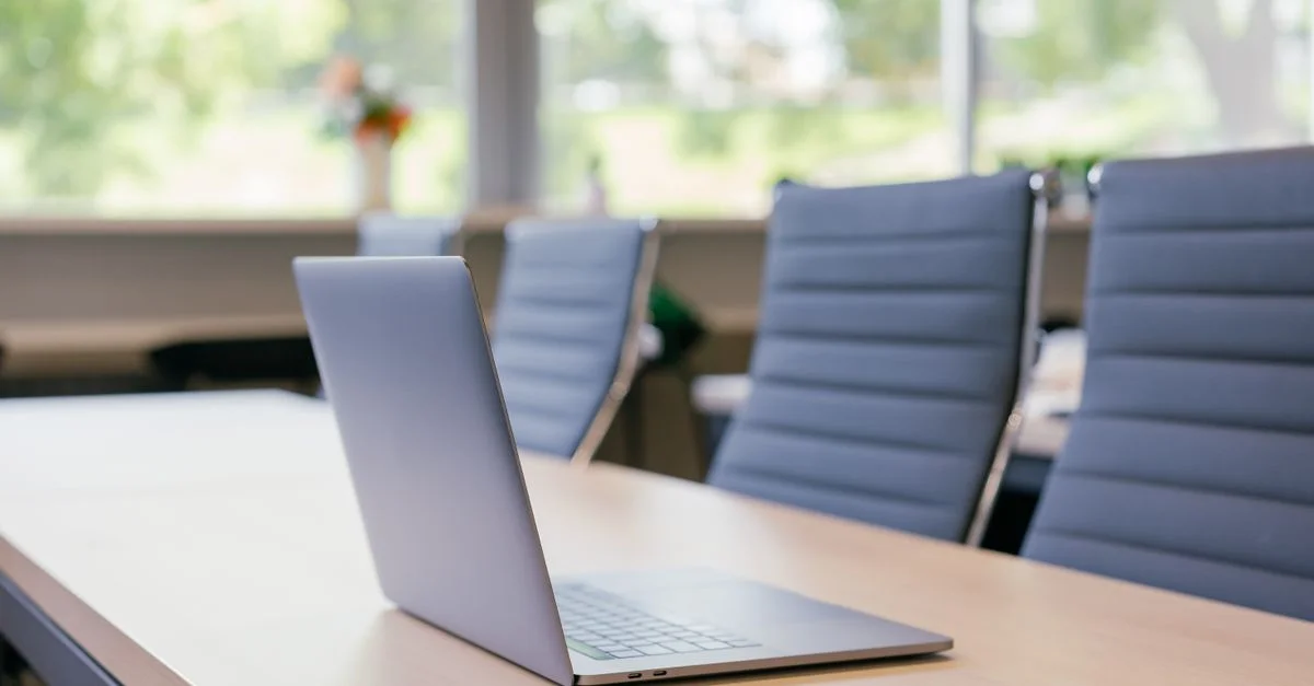 Un ordinateur portable élégant sur une table en bois dans un espace de travail de bureau ensoleillé avec des chaises de bureau bleues.