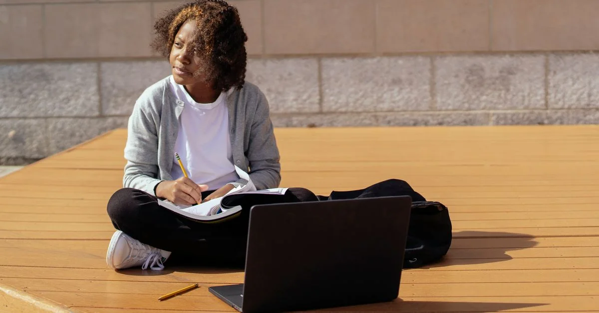 Une étudiante est assise dehors, étudiant avec un ordinateur portable et un bloc-notes sous le soleil.