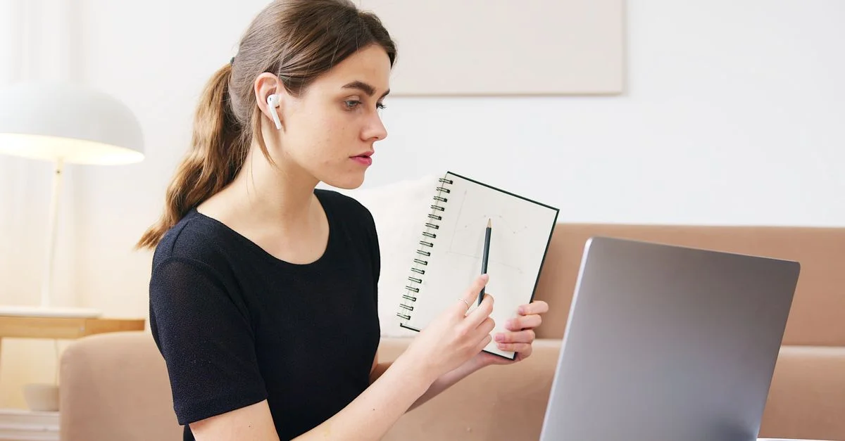 Une jeune femme lors d'un appel vidéo, prenant des notes et étudiant avec un ordinateur portable à l'intérieur.