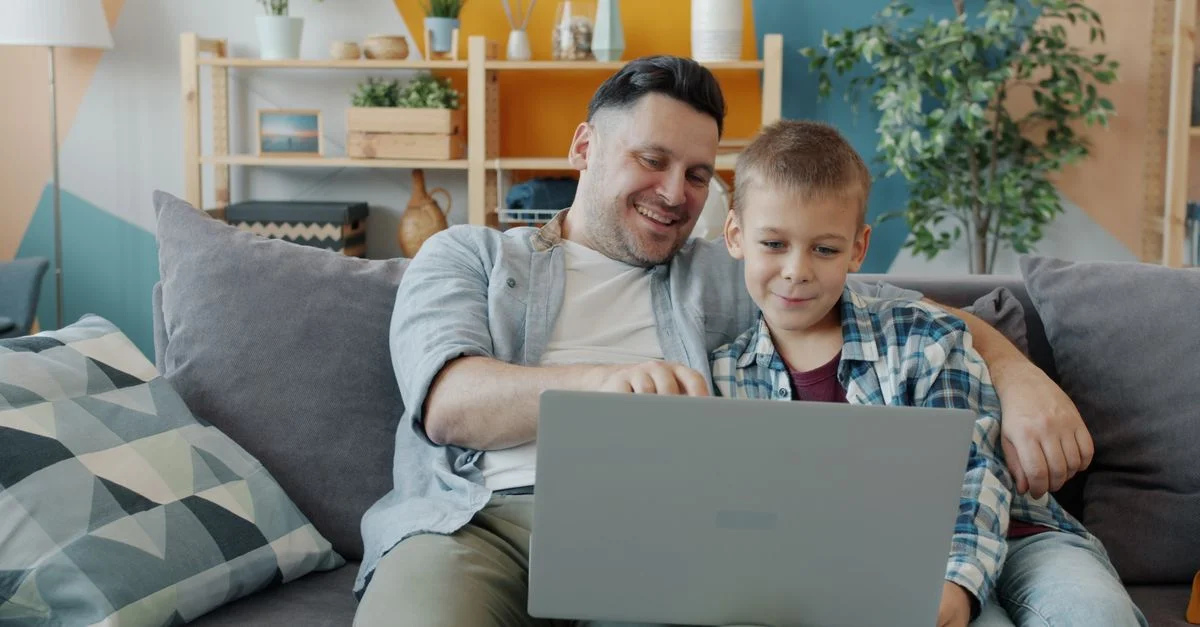 Un père et son fils, heureux, utilisent ensemble un ordinateur portable sur le canapé, profitant d'un moment en famille.