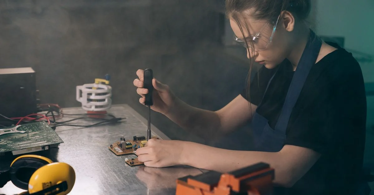 Femme jeune et concentrée dans un atelier, assemblant des circuits électroniques avec des outils de précision.