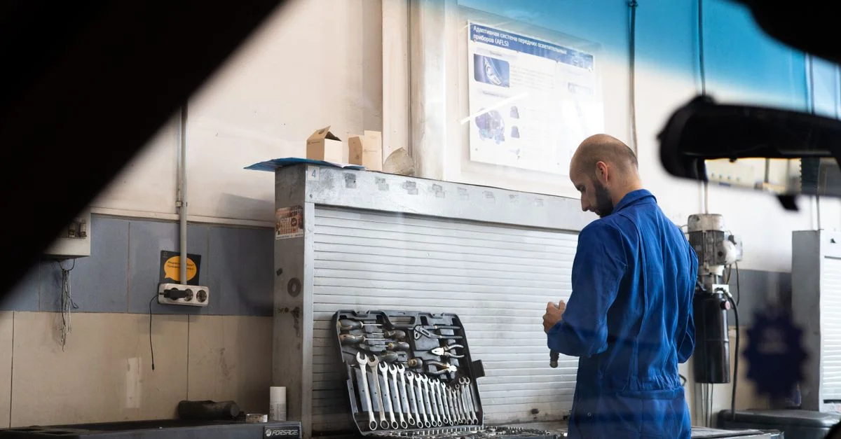 Mécanicien chauve en salopette bleue vérifiant des outils à l'intérieur d'un atelier automobile.