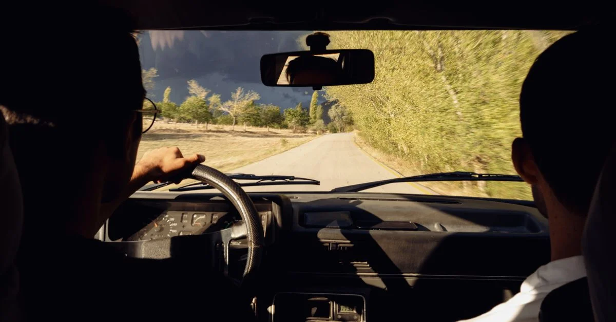 Vue de derrière de deux personnes dans une voiture circulant sur une route pittoresque, entourée d'une verdure luxuriante.