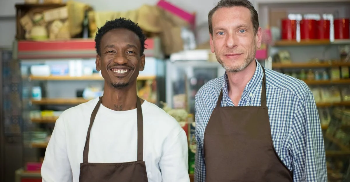 Deux propriétaires de magasin heureux souriant tout en portant des tabliers dans une épicerie locale.
