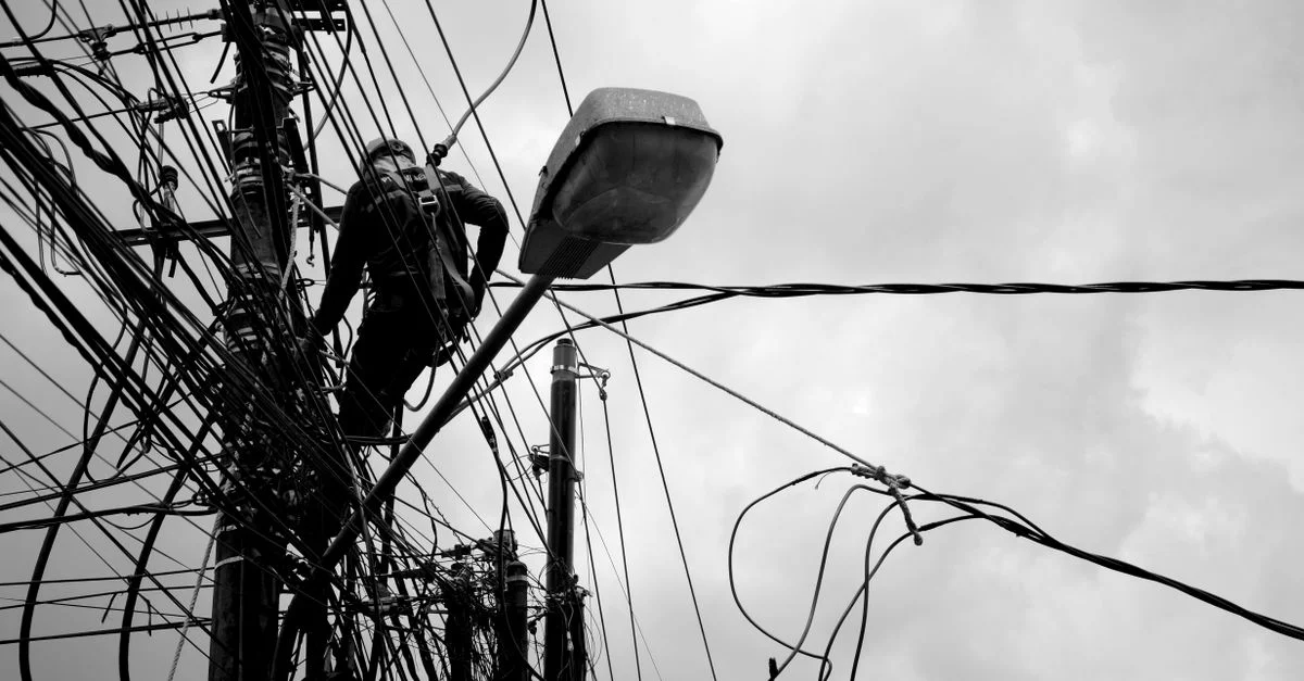 Image en noir et blanc d'un technicien travaillant sur des lignes électriques, illustrant des travaux de réparation électrique.
