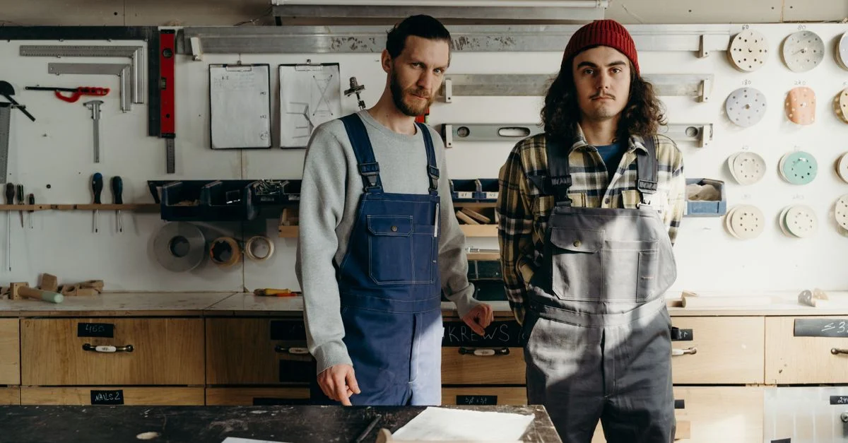 Deux menuisiers en salopette se tiennent dans un atelier bien équipé, entourés d'outils.