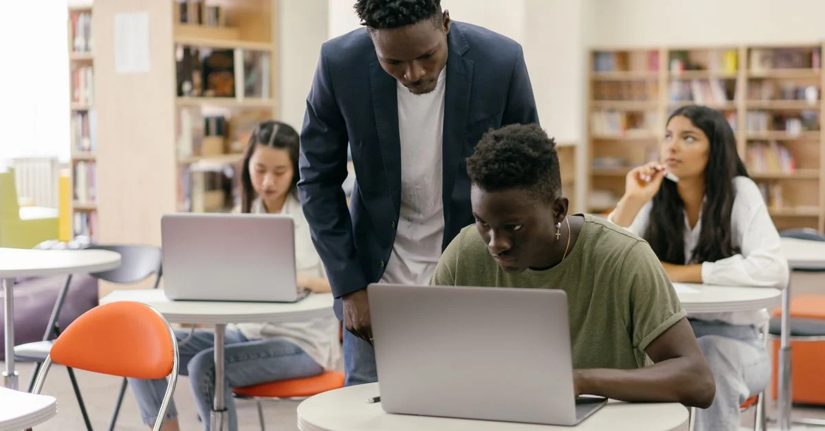 Des étudiants apprenant dans une salle de classe avec un professeur qui aide et des ordinateurs portables sur les bureaux, créant un environnement éducatif interactif.