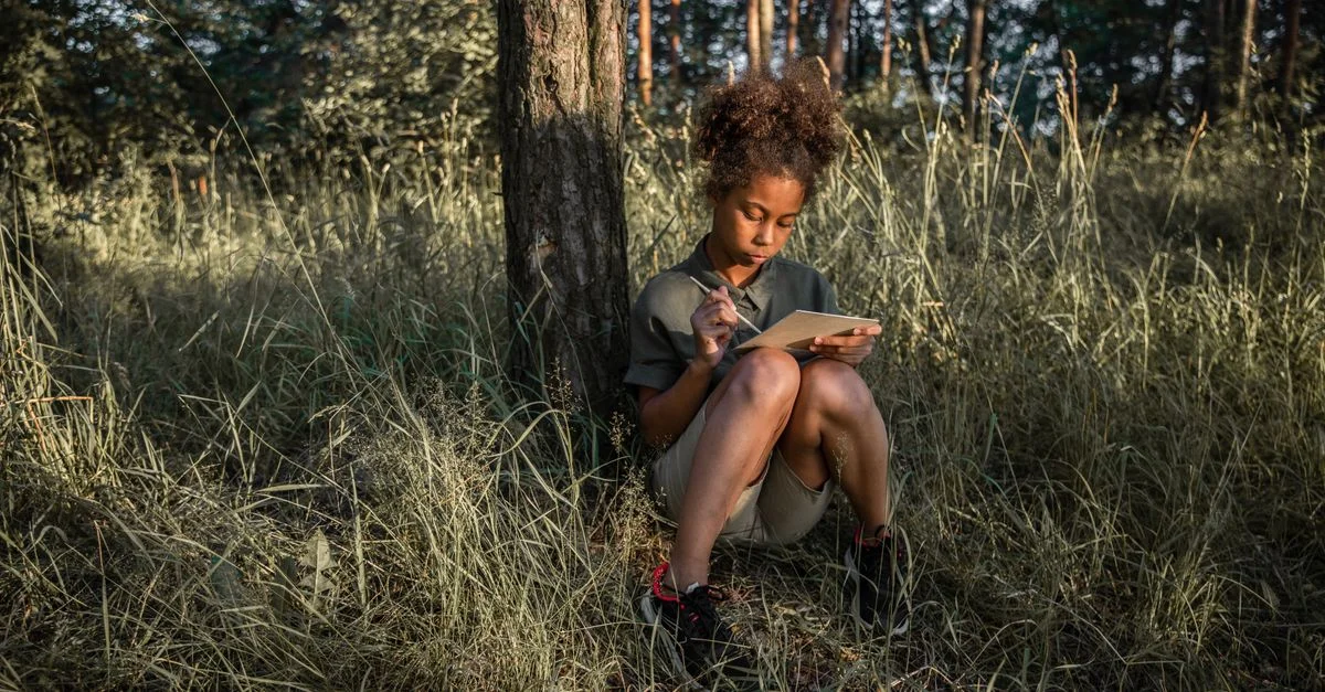 Une jeune fille aux cheveux bouclés est assise sous un arbre, dessinant ou écrivant dans un carnet de croquis dans un cadre forestier paisible.