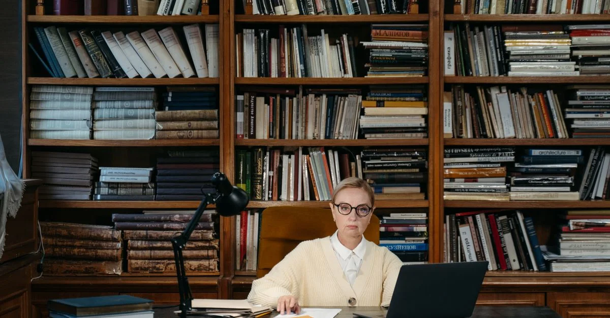 Femme concentrée travaillant sur un ordinateur portable dans une bibliothèque, entourée d'étagères.