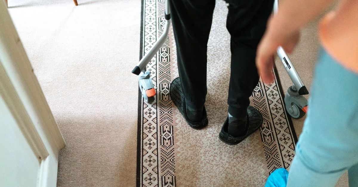 Une personne âgée avec un déambulateur assistée par un aidant dans un cadre intérieur confortable.