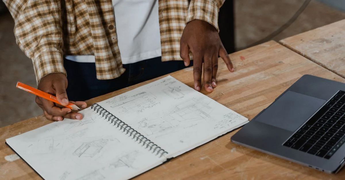 Un artisan examine des plans d'atelier détaillés, un carnet de croquis et un ordinateur portable sur l'établi.