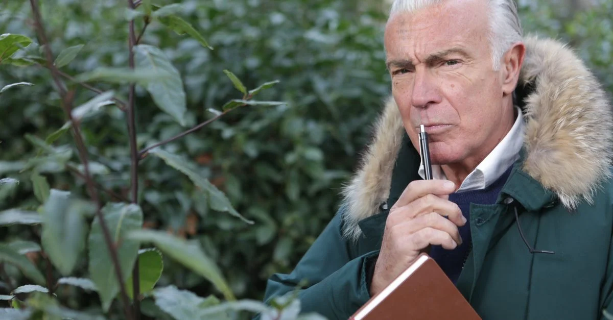 Homme âgé à l'extérieur pensant avec un stylo et un carnet au milieu de la verdure.