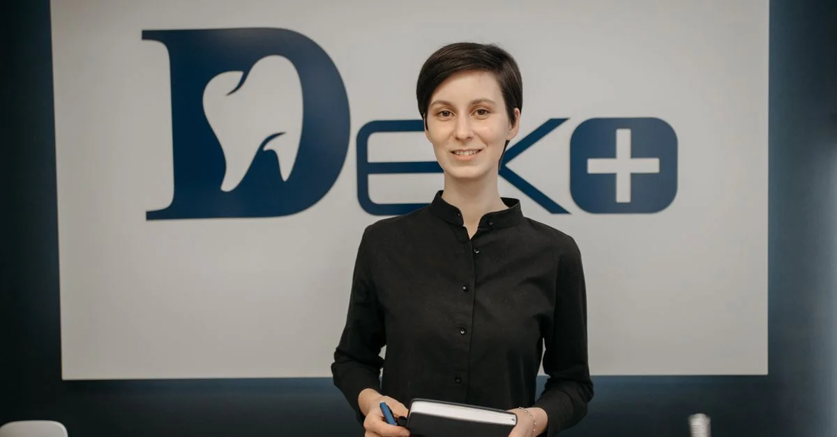 Dentiste souriante tenant un carnet devant un panneau de clinique, promouvant un environnement dentaire accueillant et professionnel.