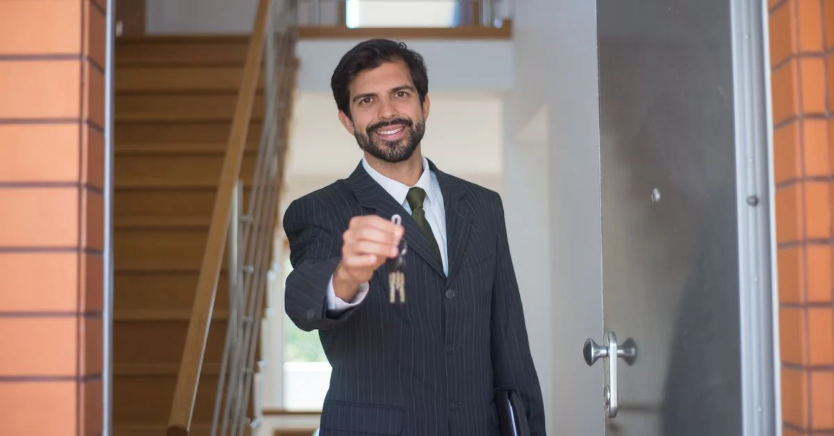 Agent immobilier souriant en costume noir remettant des clés par une porte ouverte dans une maison moderne.