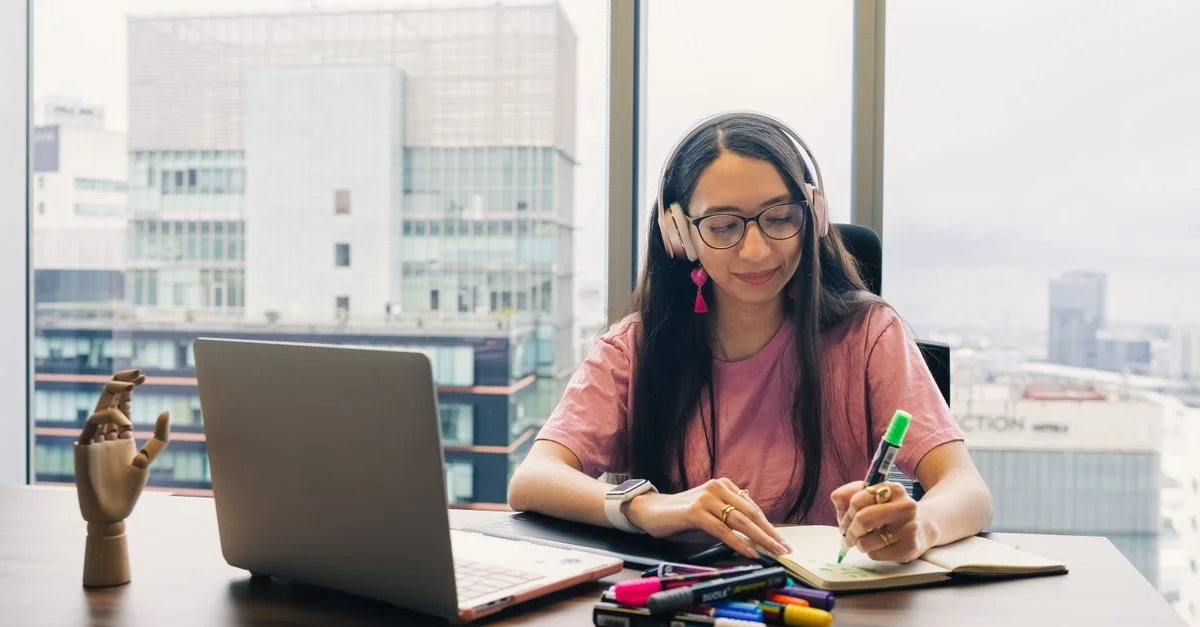 Jeune femme portant des écouteurs, écrivant dans un cahier au bureau avec vue sur la ville.