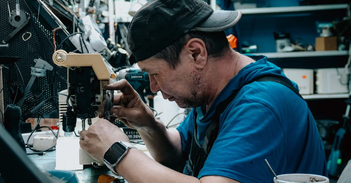 Un homme concentré, coiffé d'une casquette, répare une machine à coudre dans un atelier encombré.