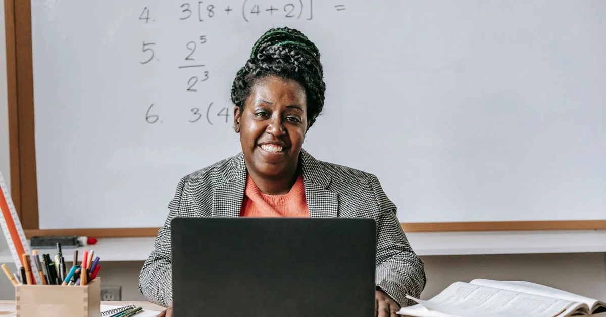Enseignante souriante utilisant un ordinateur portable dans une salle de classe avec des équations mathématiques au tableau.
