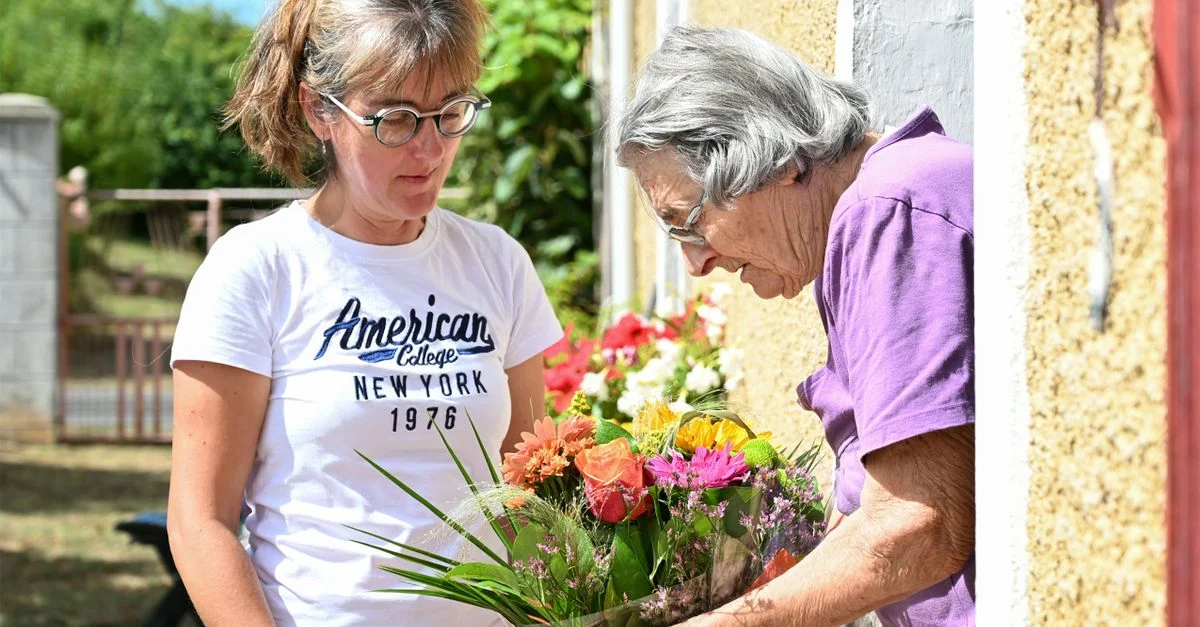 Une femme offre un bouquet vibrant à une personne âgée à l'extérieur d'une maison française.