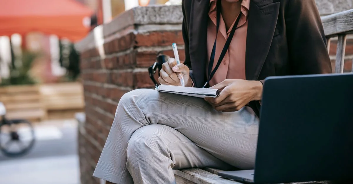 Femme d'affaires qui fait plusieurs choses à la fois à l'extérieur, prenant des notes sur un banc avec un ordinateur portable.