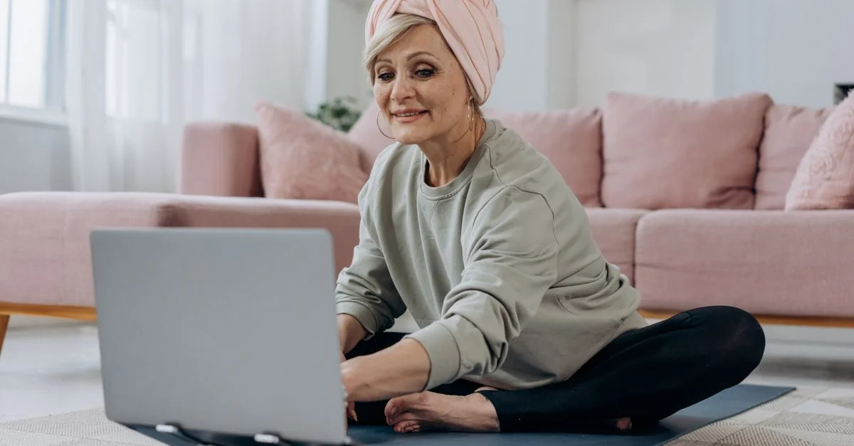 Femme senior utilisant un ordinateur portable pour un cours de yoga en ligne, assise à l'intérieur sur un tapis de yoga avec une expression détendue.