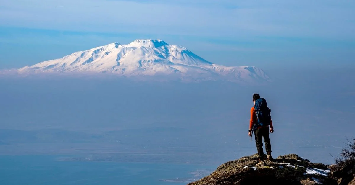 Un randonneur solitaire se tient sur une falaise rocheuse, admirant le majestueux mont Ararat enneigé.