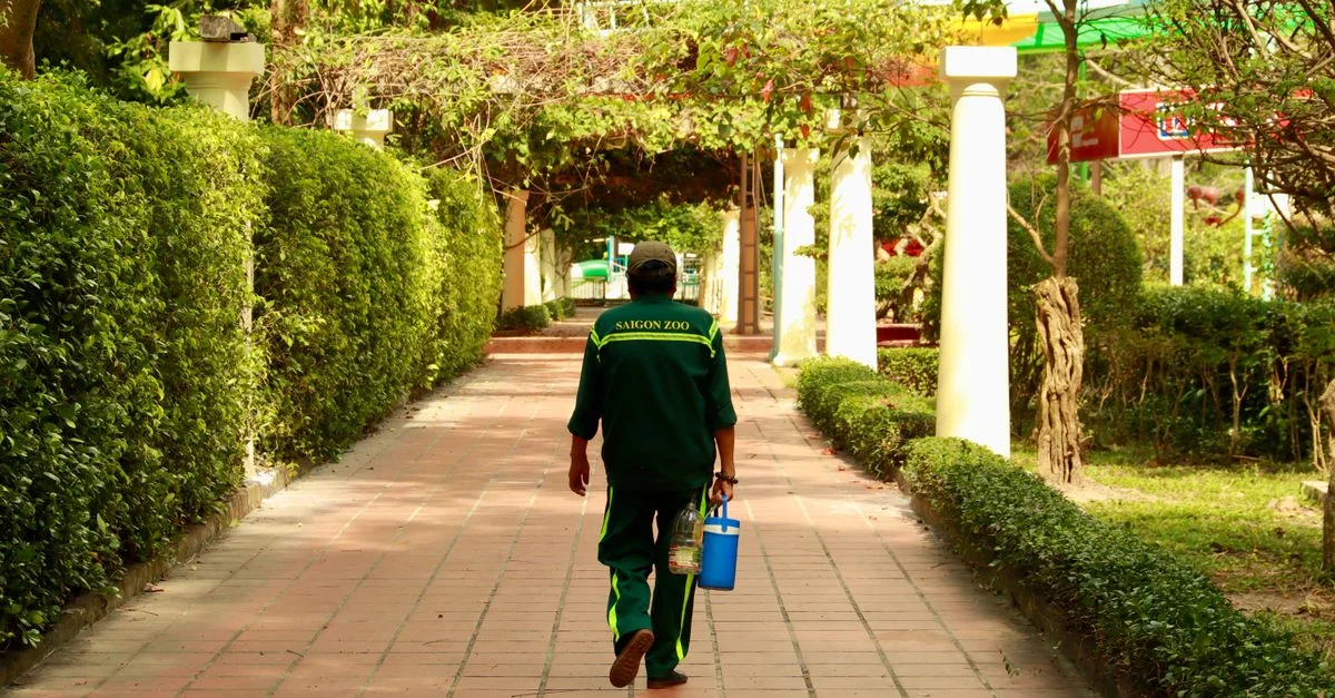 Un jardinier en uniforme se promène dans le sentier verdoyant du zoo de Saigon, portant des outils de jardinage.