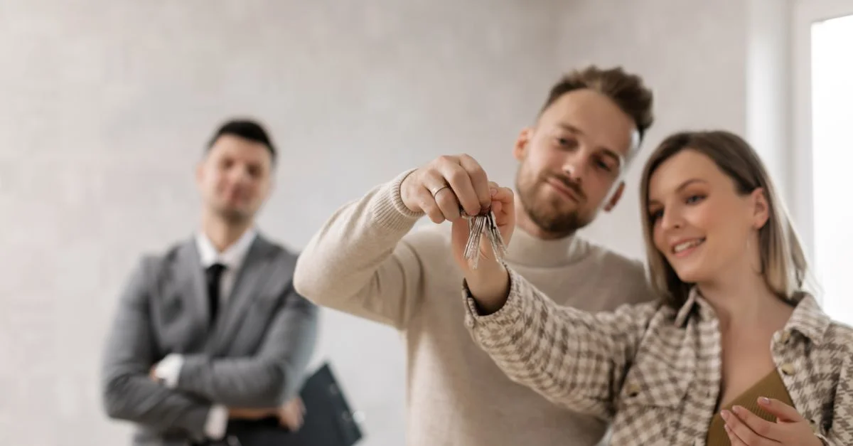 Un couple joyeux tenant les clés de leur nouvelle maison avec un agent immobilier en arrière-plan.