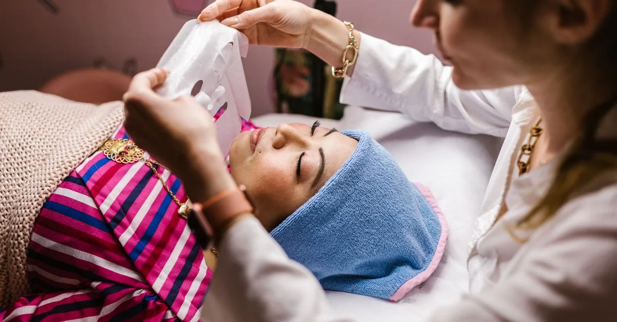 Une esthéticienne applique un masque en tissu lors d'un soin du visage pour la relaxation et le rajeunissement de la peau.