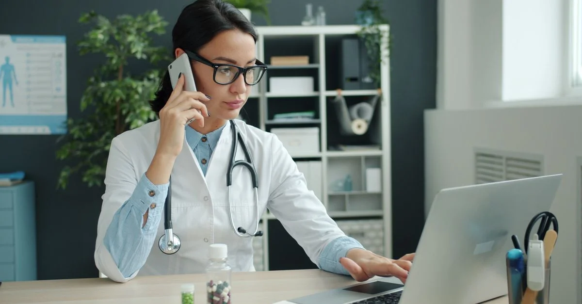 Médecin concentrée, jonglant entre son ordinateur portable et son téléphone dans un bureau moderne.