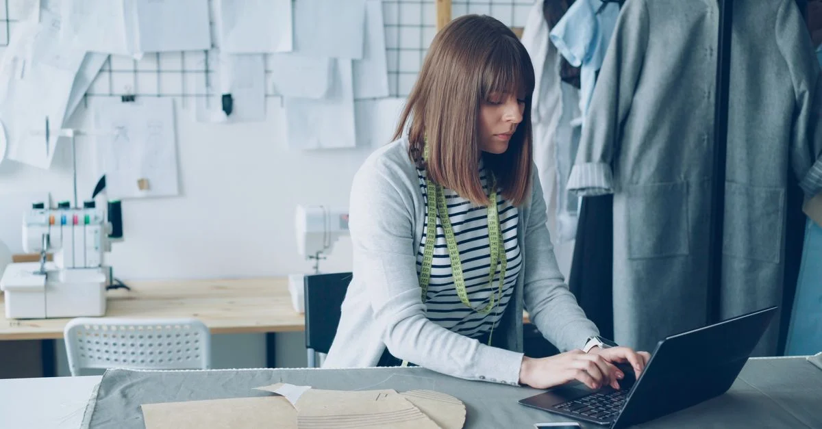 Créateur de mode travaillant sur un ordinateur portable dans un studio entouré d'outils de couture et de vêtements.