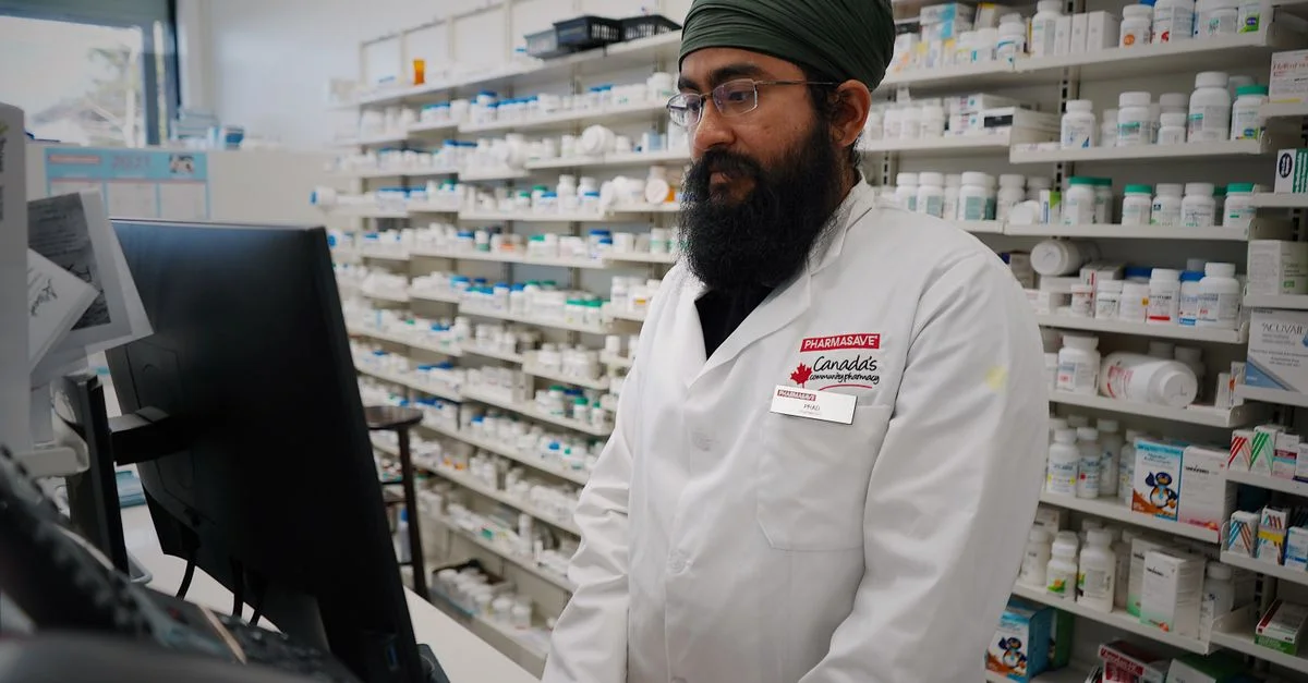 Pharmacien portant un turban travaillant sur un ordinateur dans une pharmacie avec des étagères remplies de médicaments.