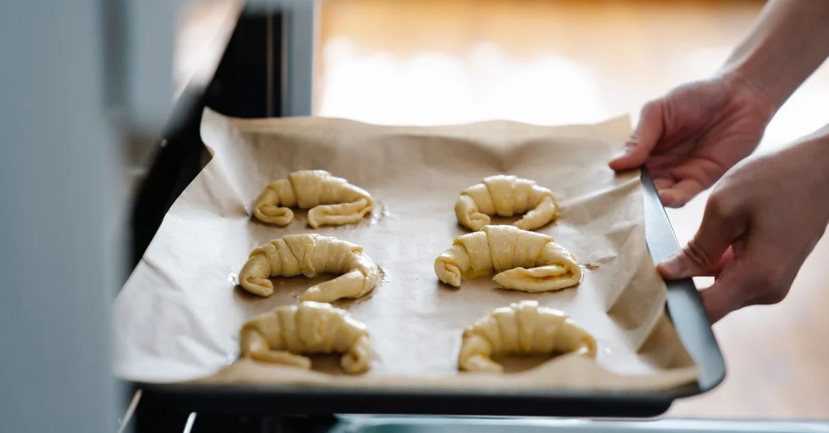 Des mains plaçant des croissants faits maison sur un plateau dans un four, prêts à cuire.