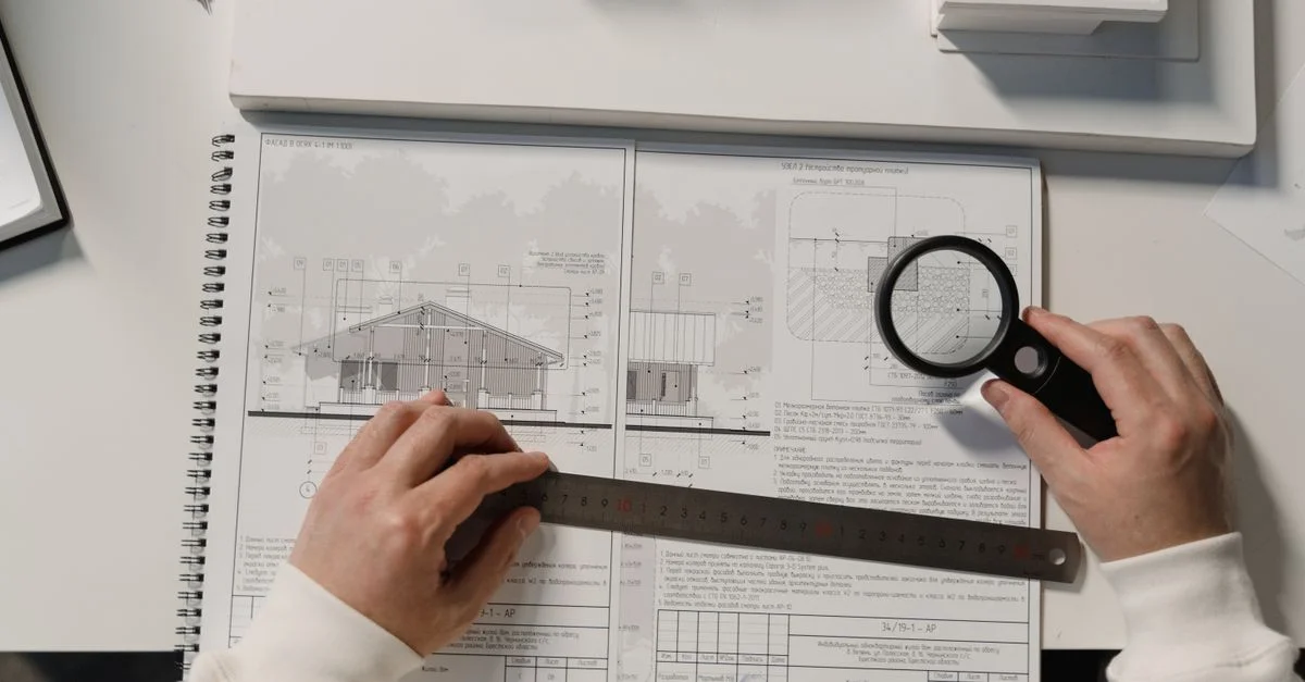 Des mains tenant une règle et une loupe sur des plans architecturaux sur une table blanche.