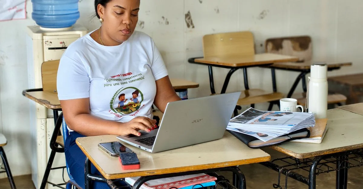 Une femme dans une salle de classe, tapant sur un ordinateur portable, entourée de matériel pédagogique, ce qui traduit sa concentration sur le travail et les études.
