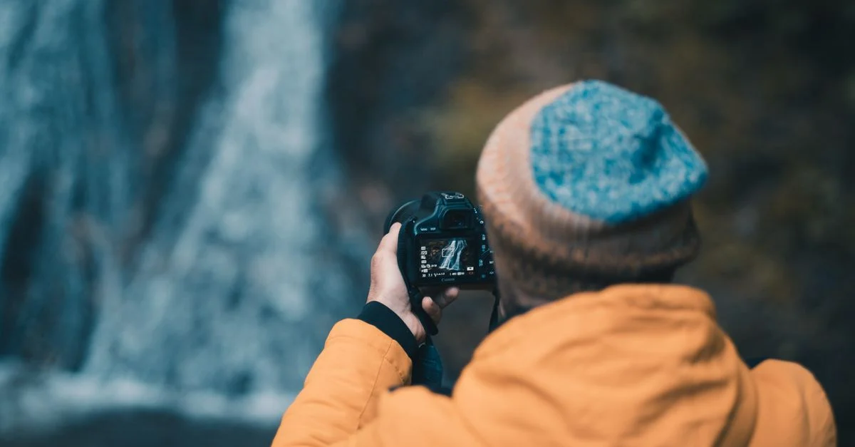 Un photographe en veste et casquette capture une cascade dans la nature.