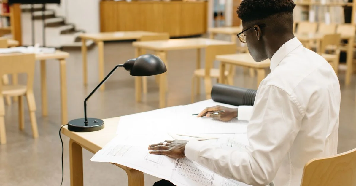 Jeune homme dessinant des plans architecturaux à une table de bibliothèque, concentré sur son travail.
