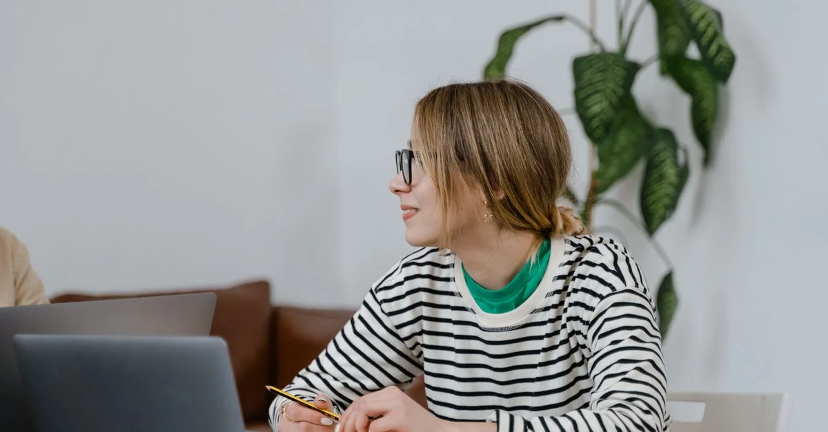 Une femme portant des lunettes est assise à un bureau, écrivant dans un carnet avec un ordinateur portable à proximité, à l'intérieur.