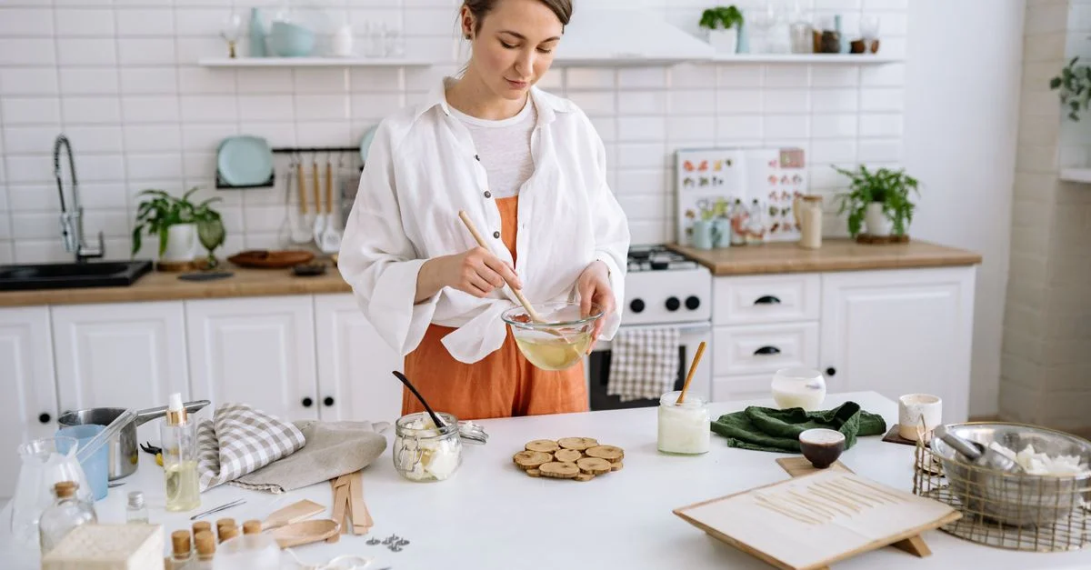 Femme remuant un bol de cire pour la fabrication de bougies dans une cuisine lumineuse et cosy.