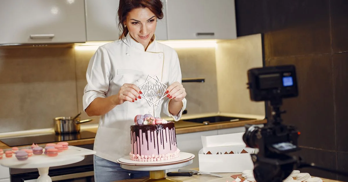 Chef femme souriante décore un gâteau tout en filmant un tutoriel de pâtisserie en ligne dans une cuisine moderne.
