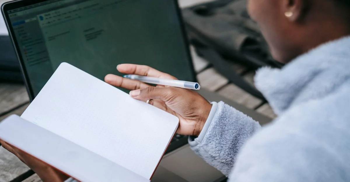 Un étudiant est assis à l'extérieur avec un ordinateur portable et un bloc-notes, concentré sur la prise de notes. Idéal pour les thèmes de l'éducation, de la technologie et du mode de vie.